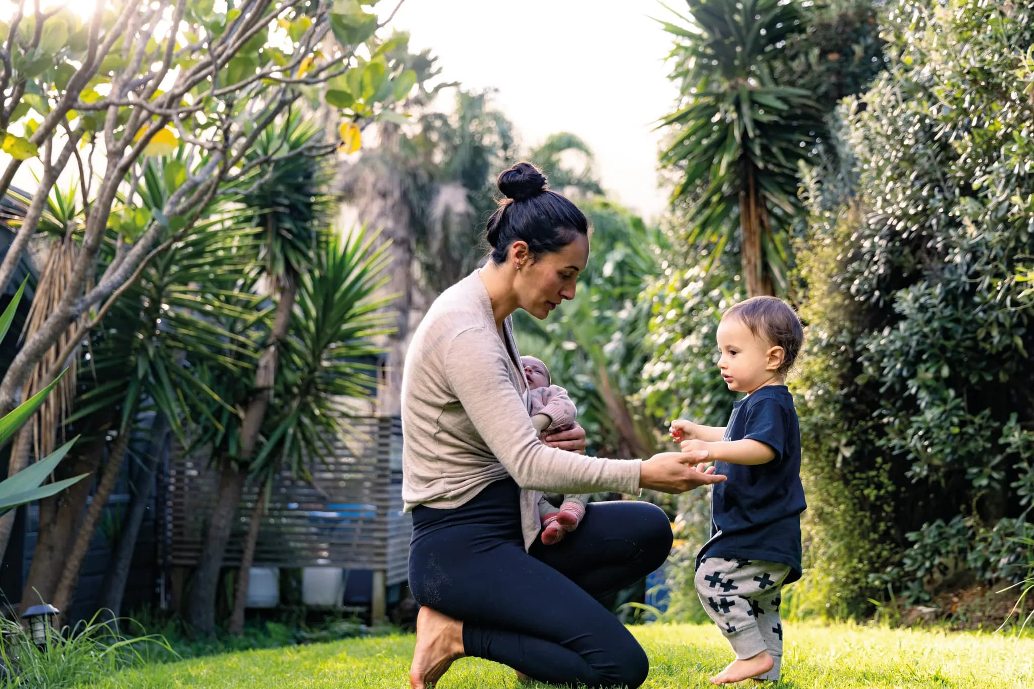 A mother holding an infant in her left arm while kneeling and holding her right hand out to a standing toddler.